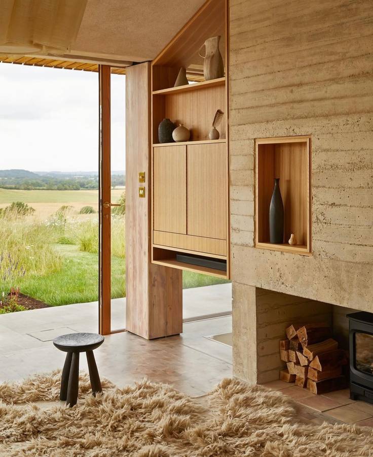 A rammed earth wall with a timber cabinet and a niche holding a vase, a fireplace with stacked firewood and a shaggy rug, with doors open to a meadow.