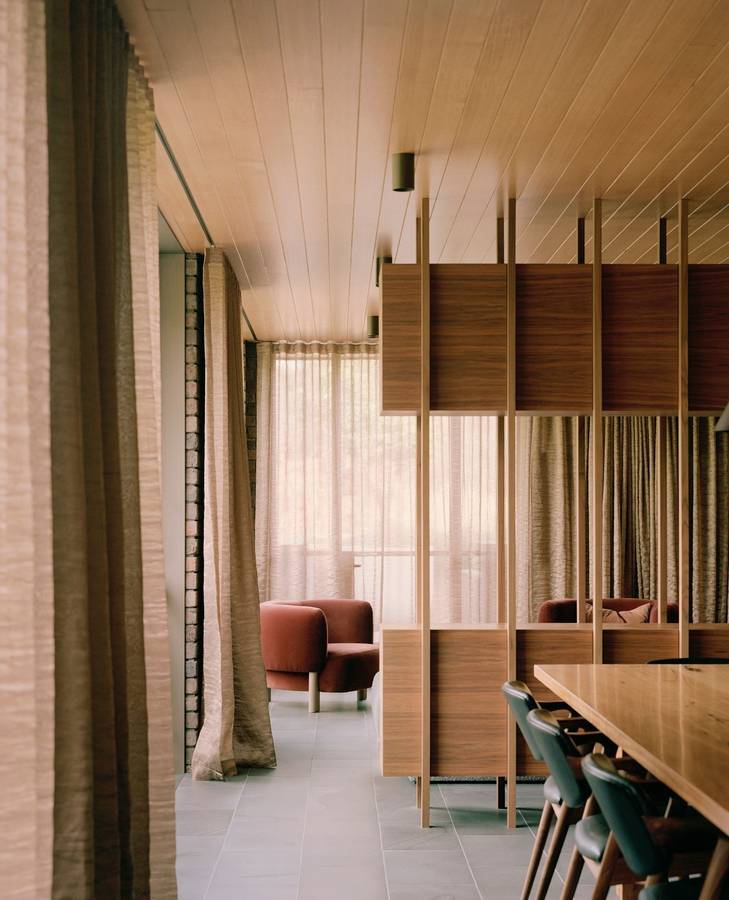A room with a timber lined ceiling and an open timber shelving screen, linen curtains, a dining table with green chairs and an armchair beyond.