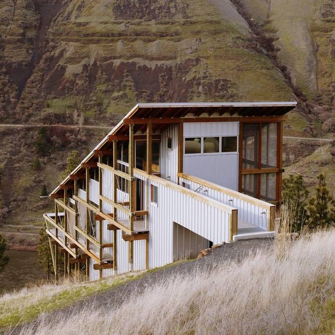 A white timber clad house on a steep hillside with an exposed timber frame and a long ramp, below layered cliffs.