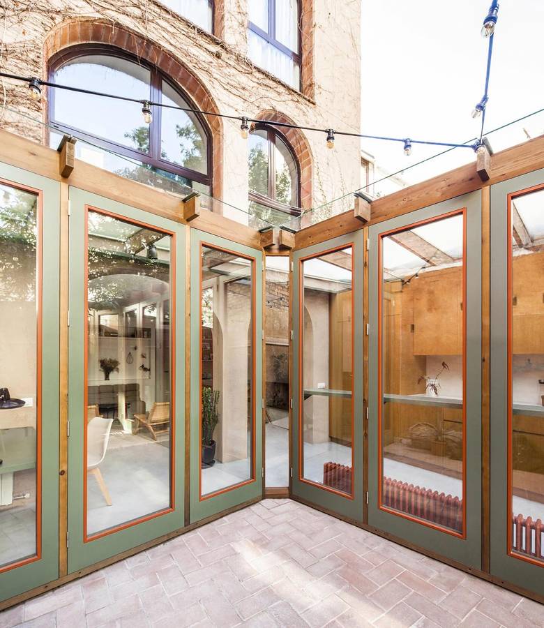 A green painted timber framed glasshouse set into the courtyard of an old brick building, with a paved floor and festoon lights.