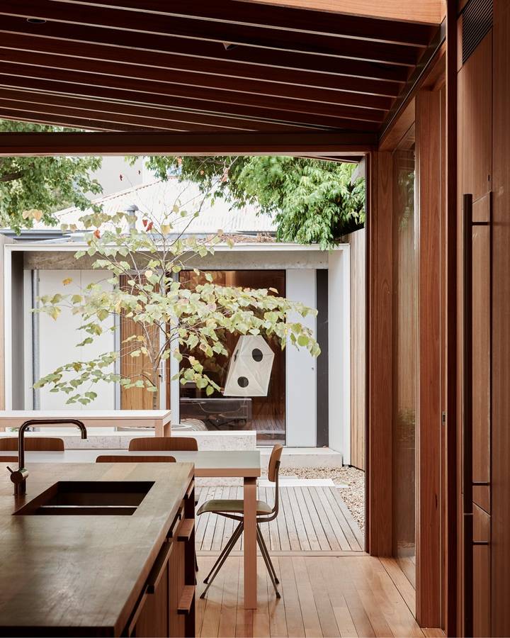 A kitchen with a dark timber ceiling and joinery, looking through sliding doors to a small courtyard with a young tree and a birdhouse.