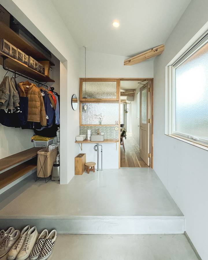 A wide concrete floored entrance hall with an open shoe store and hanging coats, a patterned glass internal window and a small hand basin.
