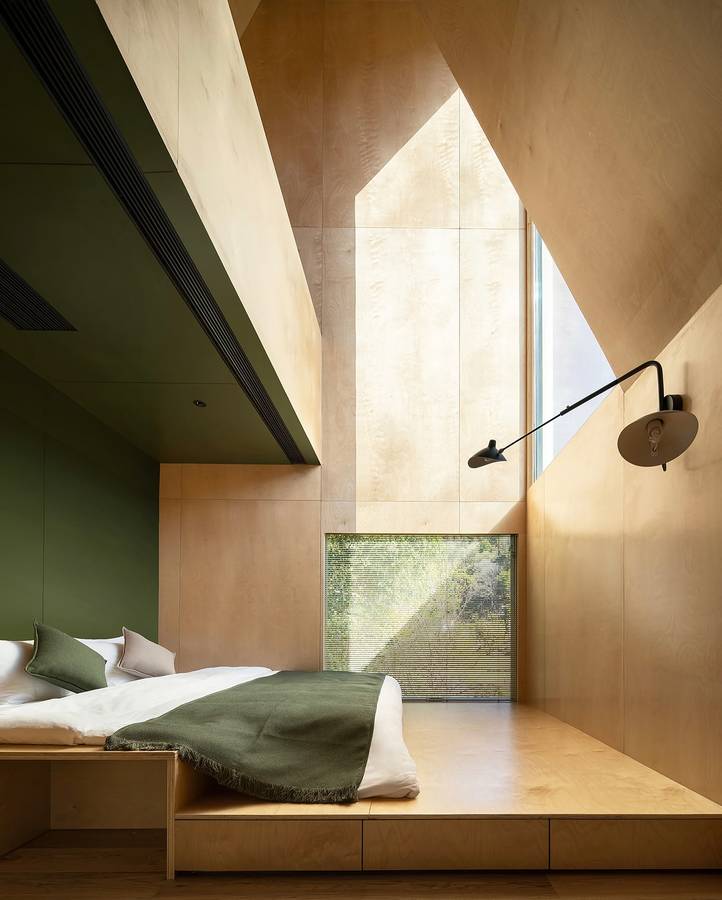 A plywood lined loft bedroom under a gabled roof with a triangular window, a green joinery wall and a platform bed.