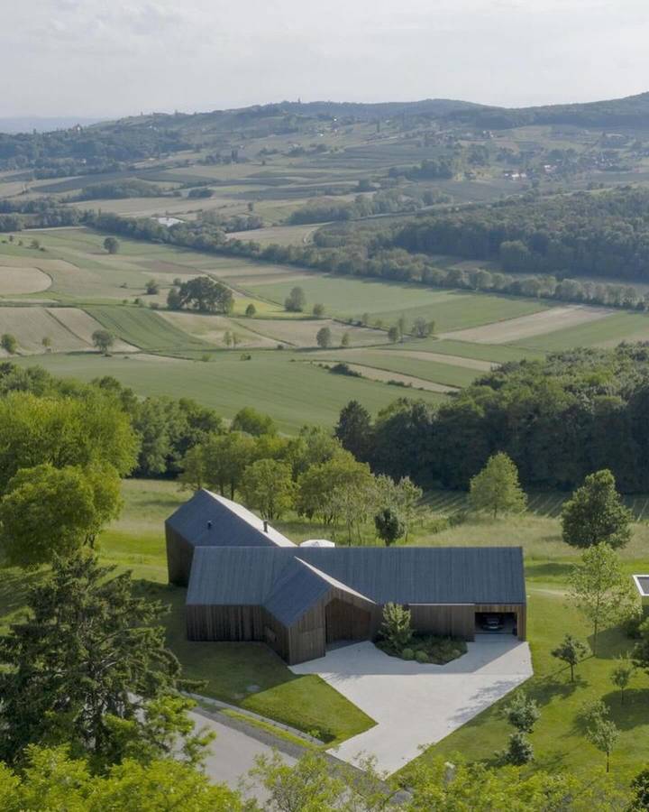 An aerial view of a dark roofed house with angled wings on a green plateau above farmland and hills.