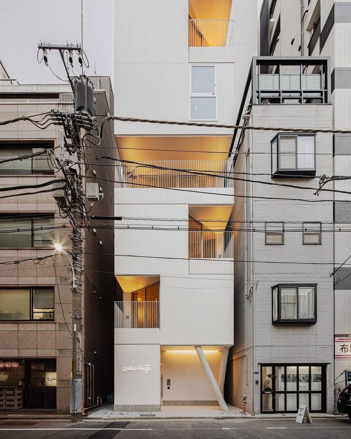 A narrow white building on a street between its neighbours, its angled window reveals lit warm at dusk under overhead power lines.