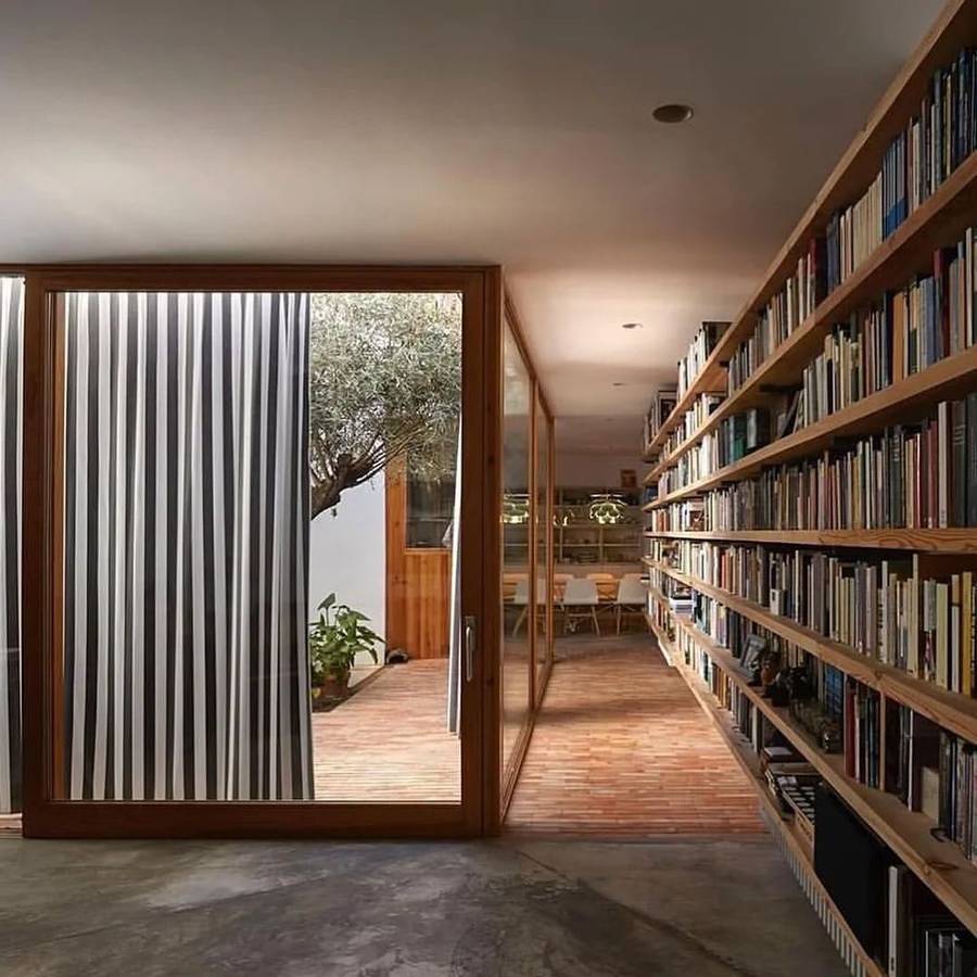 A long room with a full wall of book filled shelving on a polished concrete floor, and a glazed door to a brick courtyard with an olive tree.