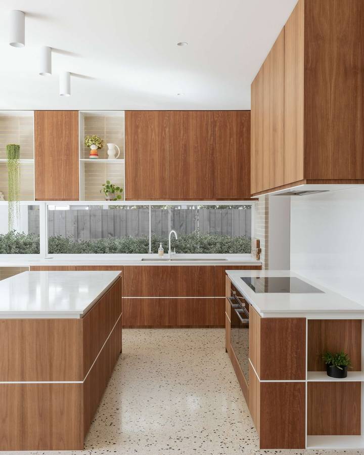 A kitchen with walnut fronted cabinets, white benchtops and a terrazzo floor, with a long window over the sink.