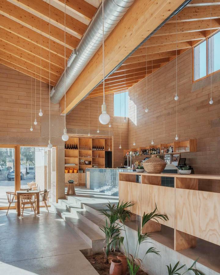 A double height room with an exposed timber roof structure and ducting, plywood lined walls, a bar counter and a stepped concrete floor with planting.