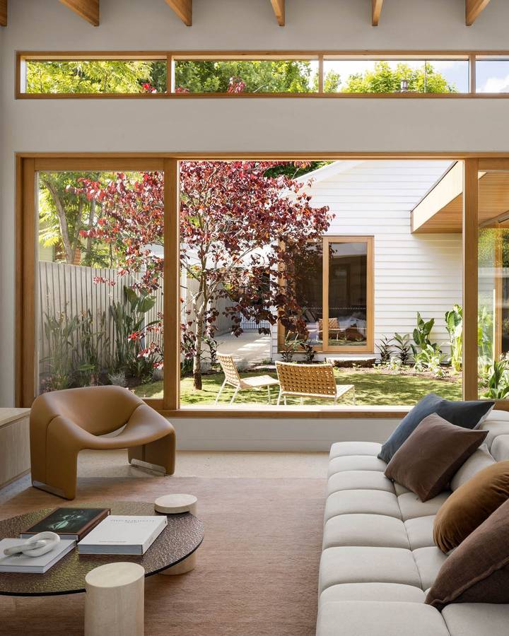 A living room with clerestory windows and a large picture window to a courtyard with a red leaved tree, a pale sofa and a tan armchair.