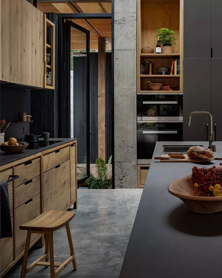A kitchen with blackened timber and oak joinery against a board formed concrete wall, with a dark benchtop and a timber stool.