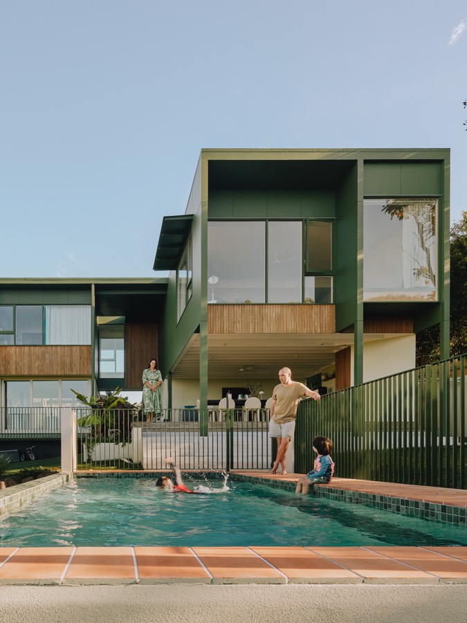A two-storey house clad in green, with a pool in front and a family around it.