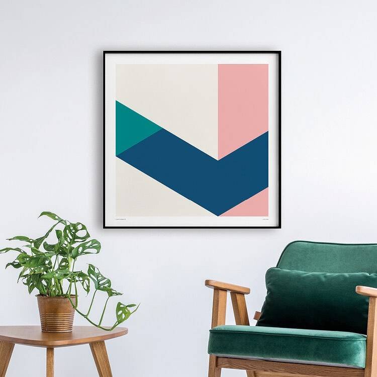 A framed geometric print in teal, navy, pink and cream hung above a plant and a green velvet armchair.