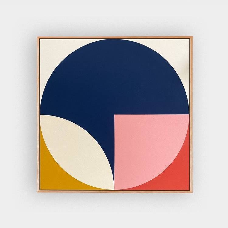 A square canvas in a pale timber frame painted with a navy circle, a pink square and mustard, cream and red segments.