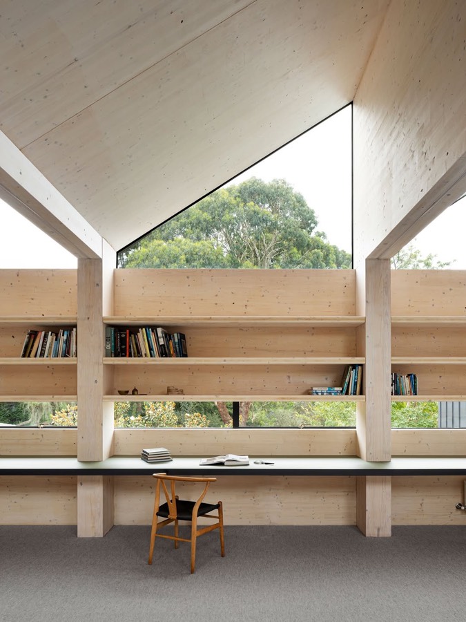 A room lined in pale timber with a long built-in desk, bookshelves and a triangular window under the roof.
