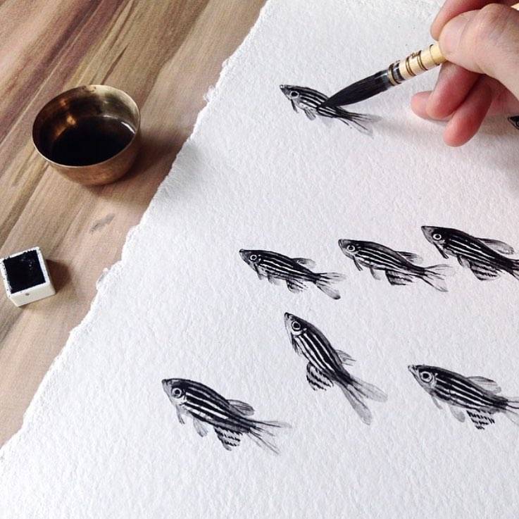 A hand painting rows of striped fish in black ink on textured paper, beside a brass bowl of ink.