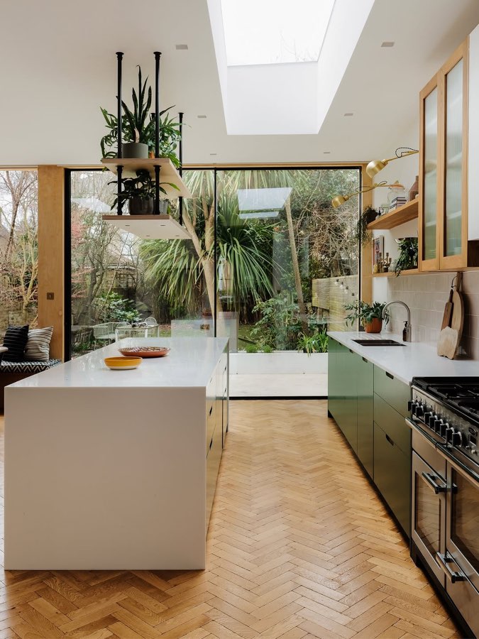 A kitchen with a long island, a herringbone parquet floor and a skylight, opening to a green garden.