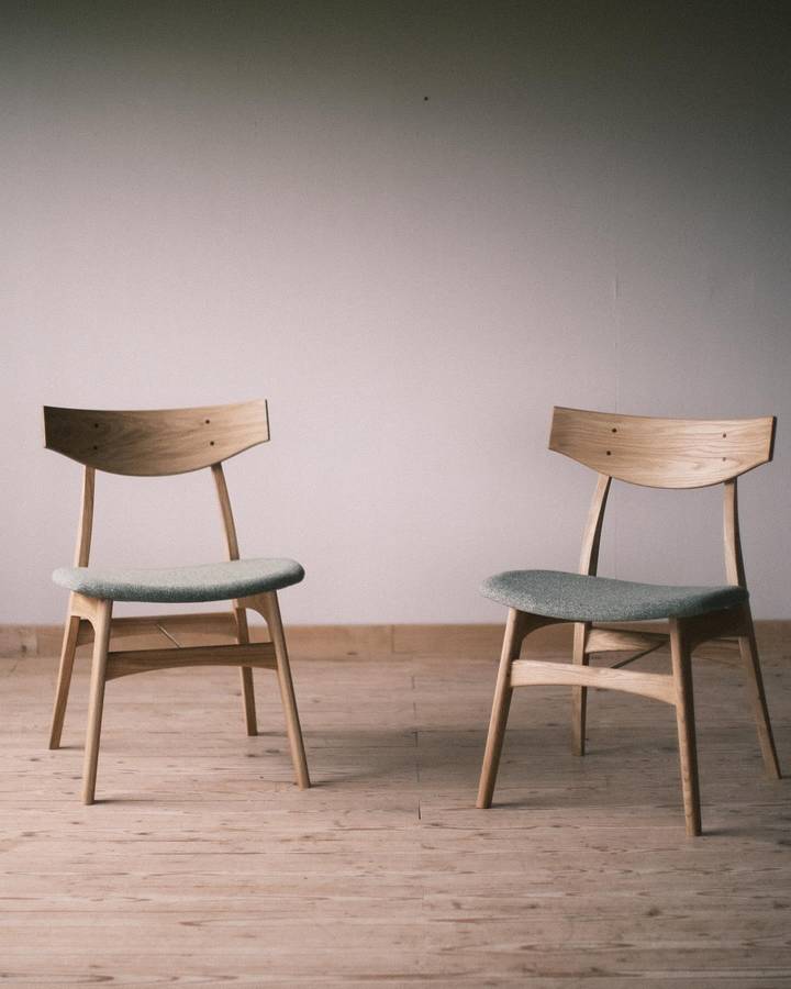 Two oak dining chairs with curved backrests and green upholstered seats, standing apart on floorboards against a pale wall.