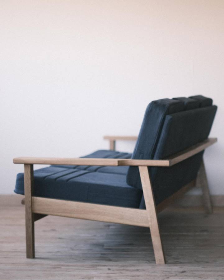 A three seater sofa with an oak frame and dark blue cushions, seen from behind with a timber rail running from the arm around the back.