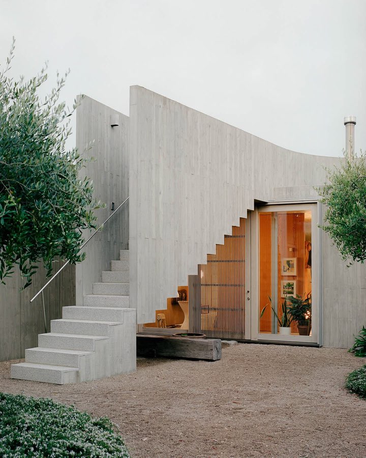 A concrete house with a curved wall, an external stair and an open timber door, with olive trees beside it.
