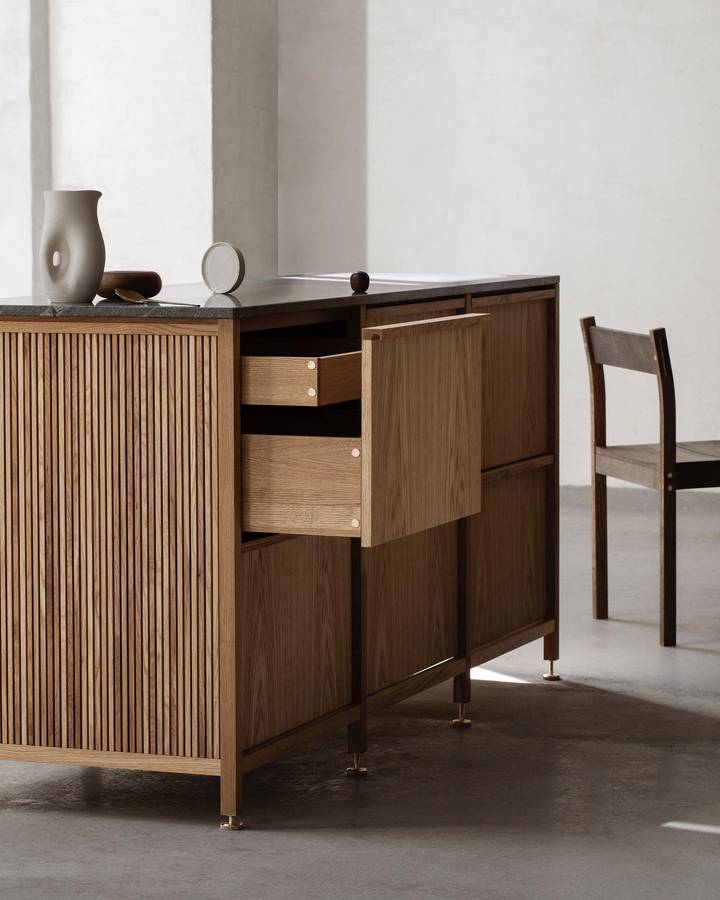A freestanding oak kitchen island with a fluted end panel, a dark stone benchtop and two drawers pulled open, on a concrete floor.