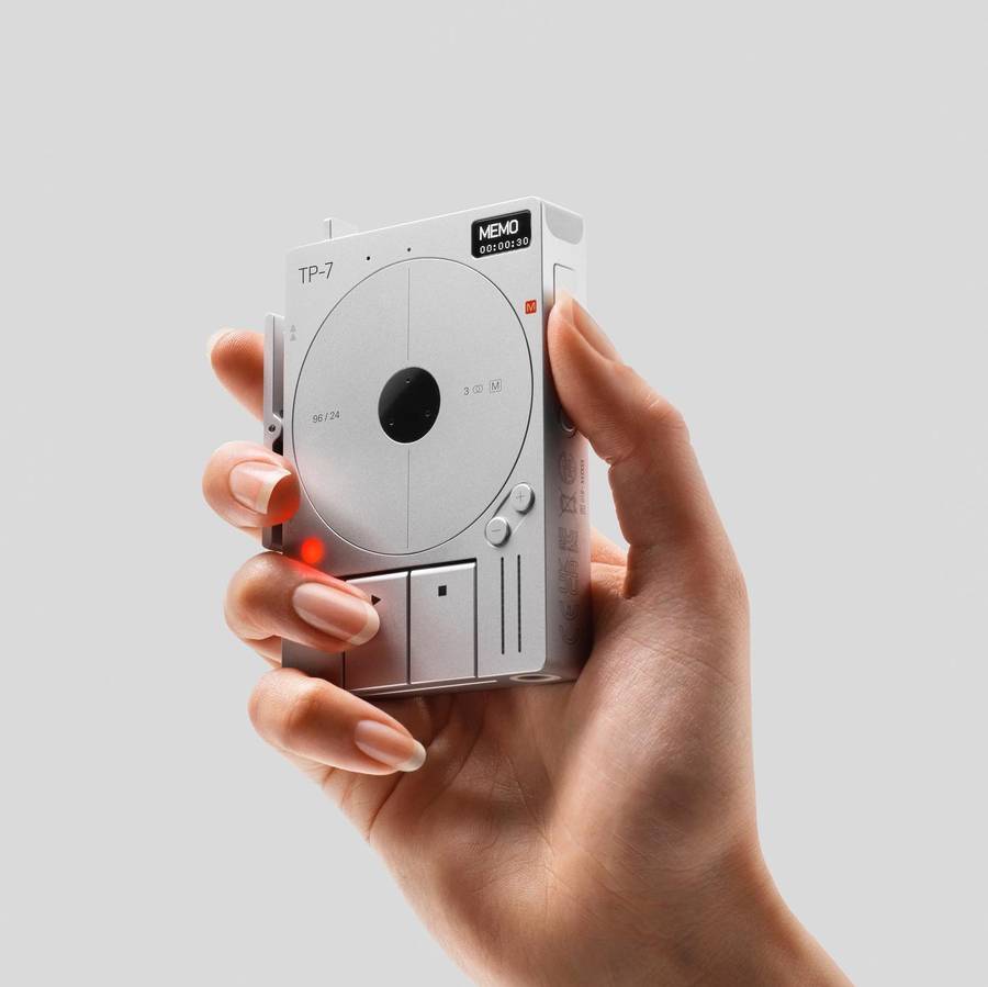 A hand holding a small silver recorder with a circular tape reel and a screen reading MEMO, on a plain pale grey ground.