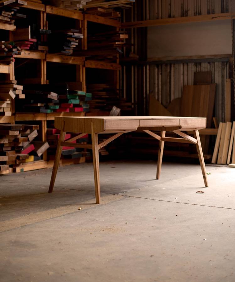 A pale timber desk with two drawers on splayed legs, standing on a concrete floor in a workshop with racks of timber behind.