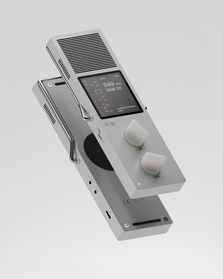 A grey portable radio with two conical knobs, a wire handle and a screen reading 549 kHz, tilted against a plain grey ground.