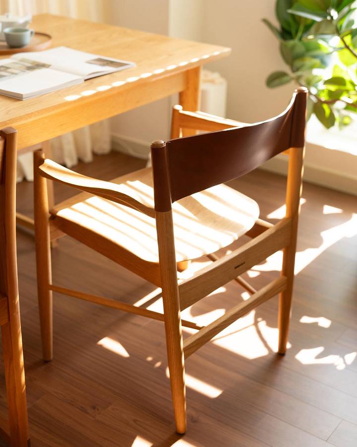 A pale timber armchair with a dark brown leather backrest beside a timber dining table, in sunlight on a wood floor.