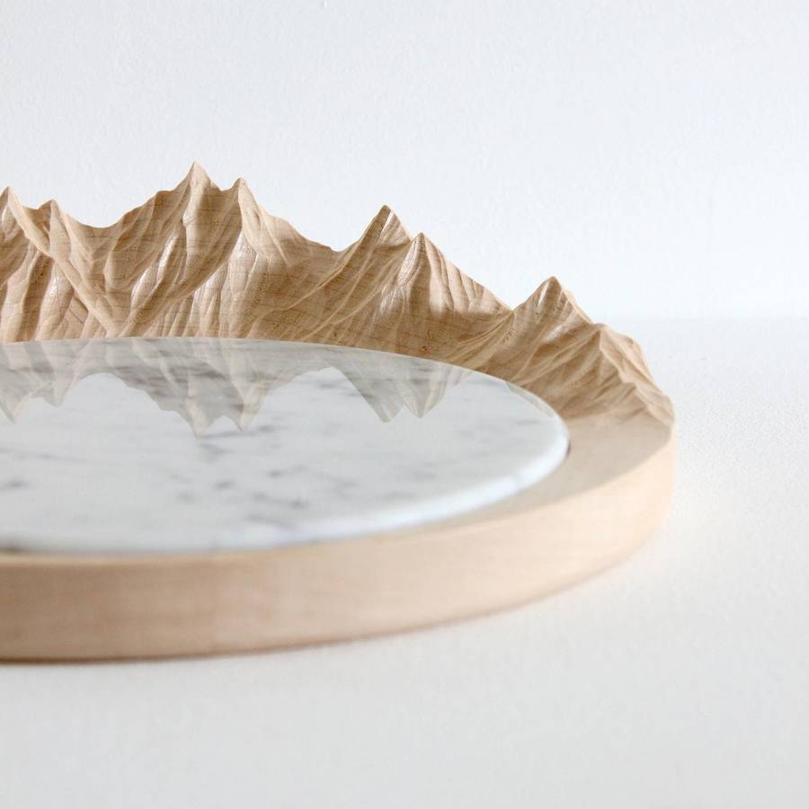 A round pale timber board with a carved mountain range along one edge and a white marble inset in the centre.