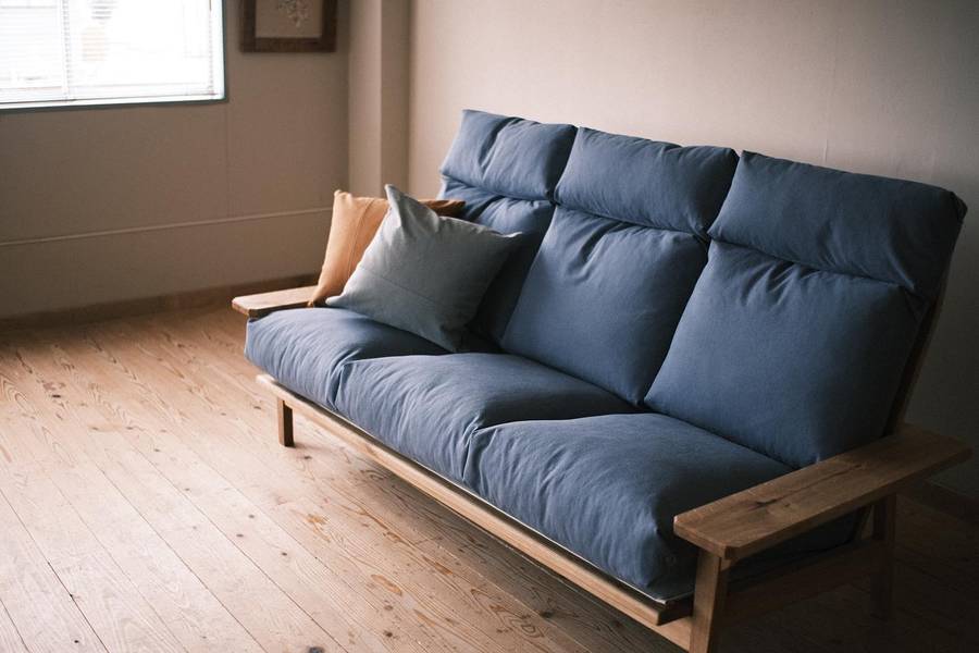 A three seater sofa with an oak frame, a high back and grey blue canvas cushions, standing on floorboards with two square cushions on the seat.