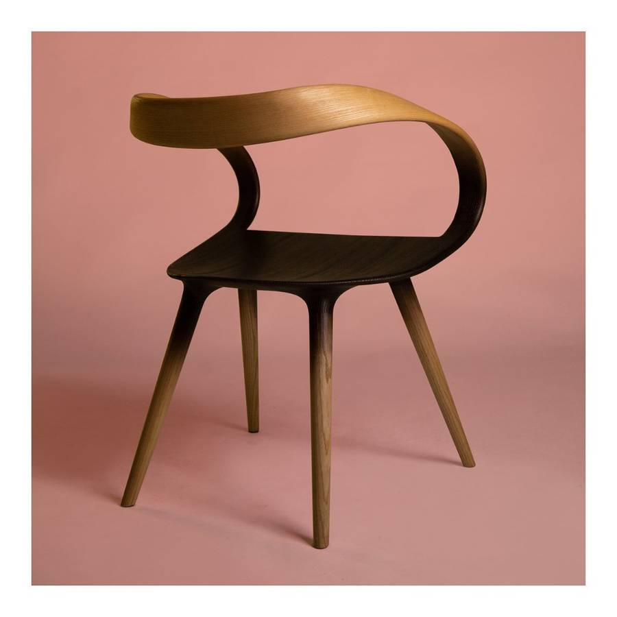 A timber chair with a curved one piece back and arm, its colour fading dark through the seat and arm loop, on a pink ground within a white border.