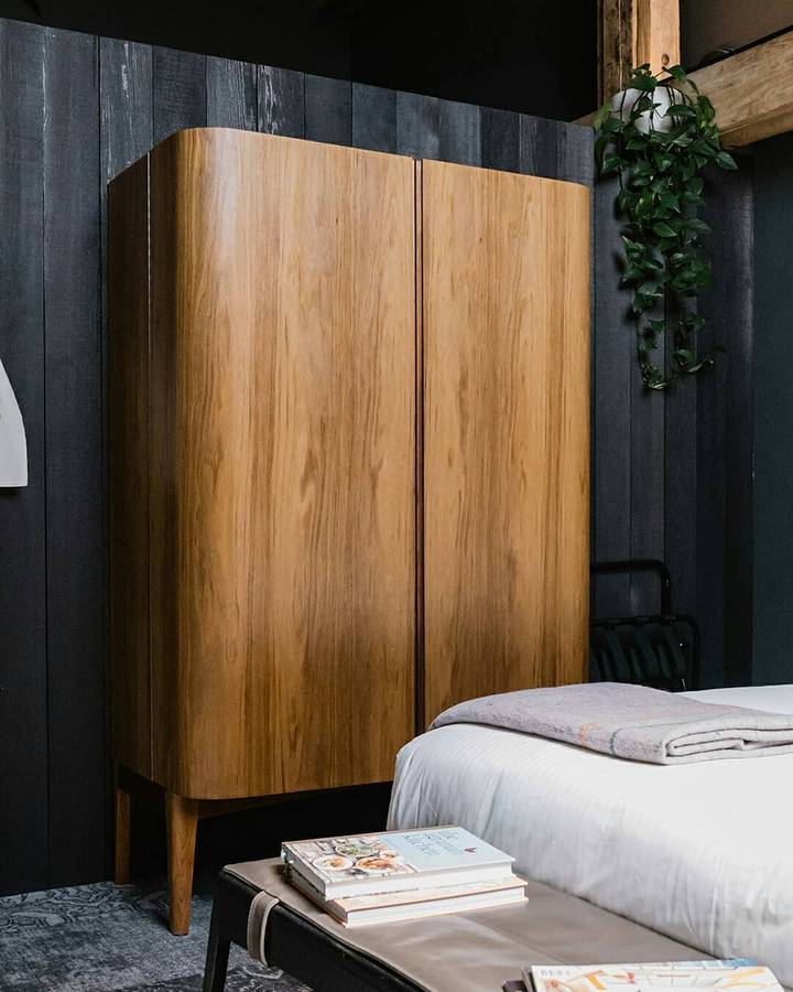 A tall two door timber cabinet with rounded corners on tapered legs, standing against a black timber board wall beside a bed, with a bench of books and a hanging plant.