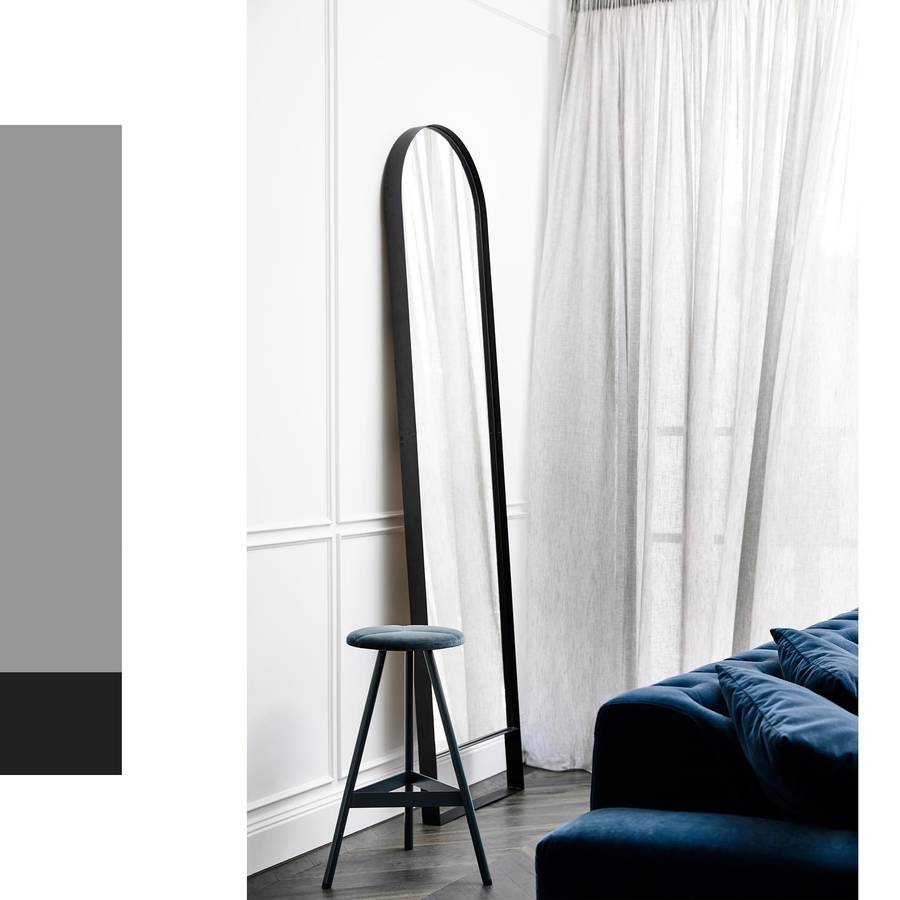 A tall black framed arch mirror leaning against a white panelled wall beside sheer curtains, with a tripod stool in front and a blue velvet sofa at the right, and a grey and black colour block down the left edge.