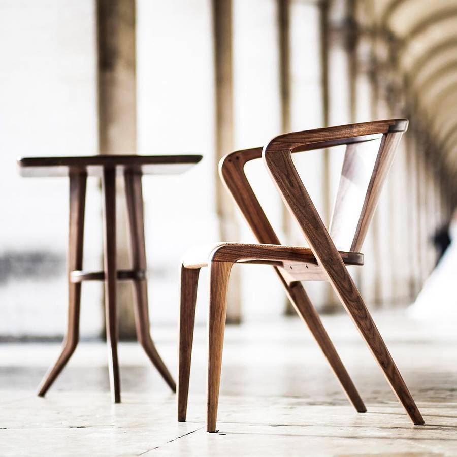 A dark timber armchair with splayed legs and a pale seat, beside a small timber side table on a stone floor in a colonnade.