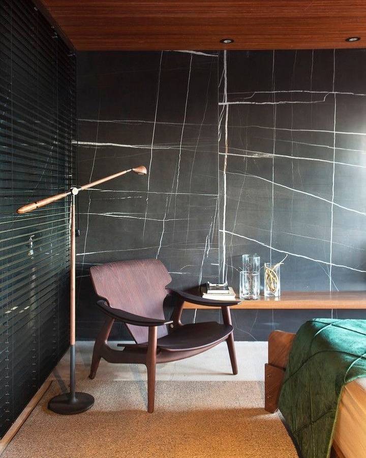 A bedroom corner with a black marble wall and a timber ceiling, a timber lounge chair, a copper floor lamp and a floating timber shelf holding two glasses and a book.