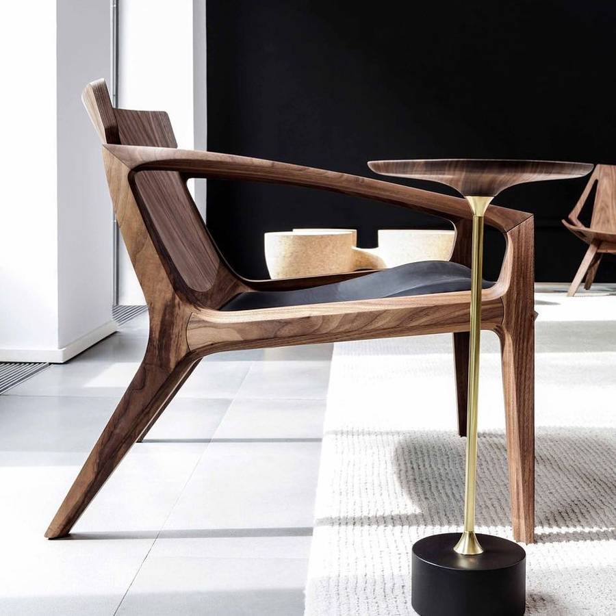 A walnut lounge chair with a swept frame and a black leather seat, beside a slim side table with a timber top on a brass stem and black base, on a pale tiled floor.