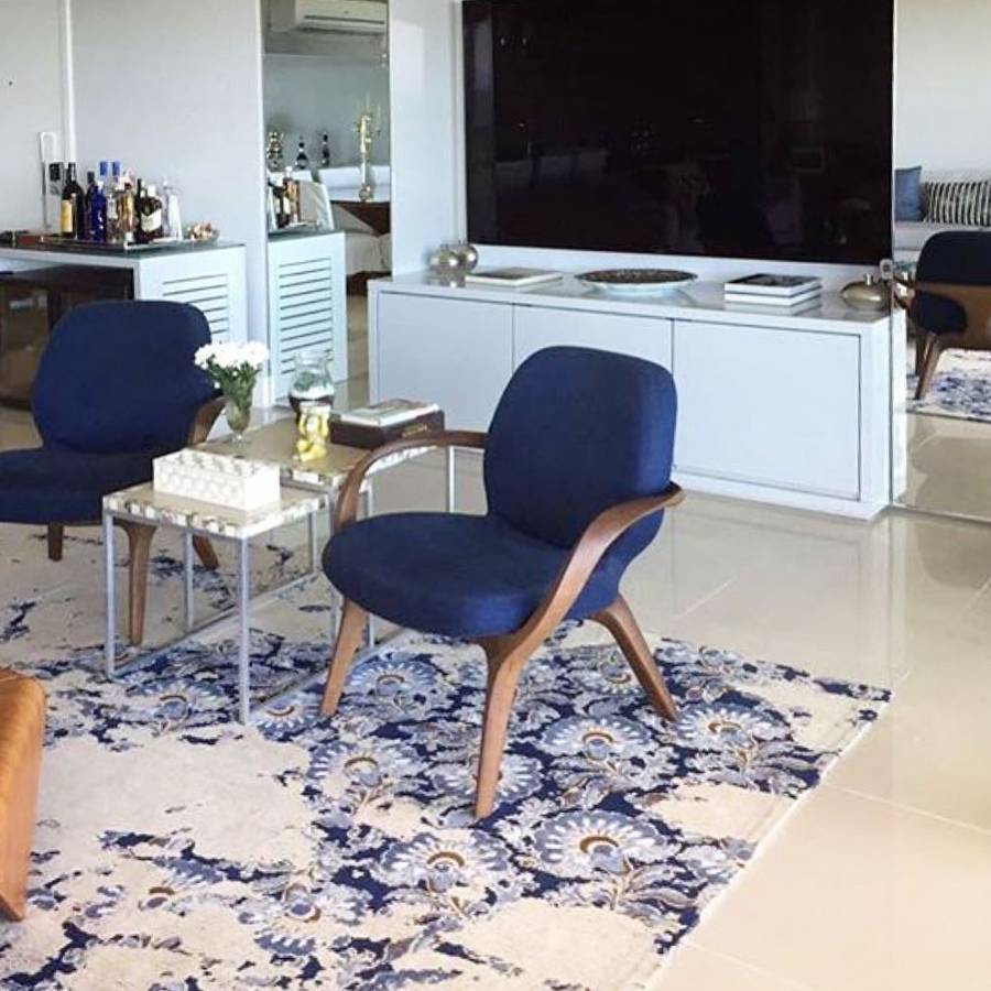 A living room with two blue upholstered armchairs on timber frames, a floral rug, a glass side table, a white sideboard and a large dark window.