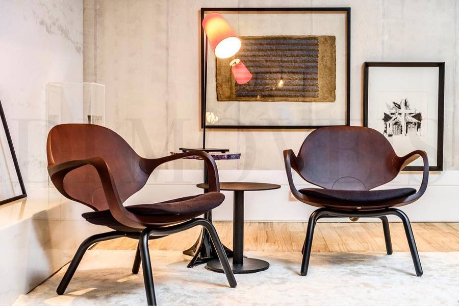 Two timber shell lounge chairs with black metal legs facing each other across a small round table, with a floor lamp, two framed works and a pale rug in a concrete walled room.
