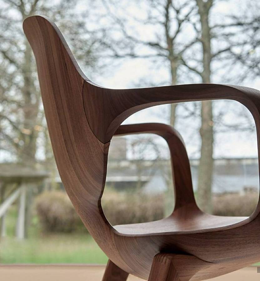 A close view of a solid timber chair, showing the swept back rail meeting a looped arm, with bare trees and a lawn blurred behind.