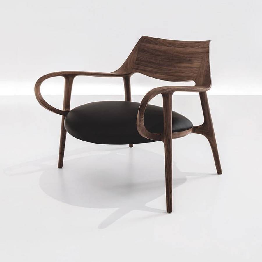 A walnut lounge chair on a white ground, with a wide curved back shell, looped arms, slim tapered legs and an oval black leather seat.