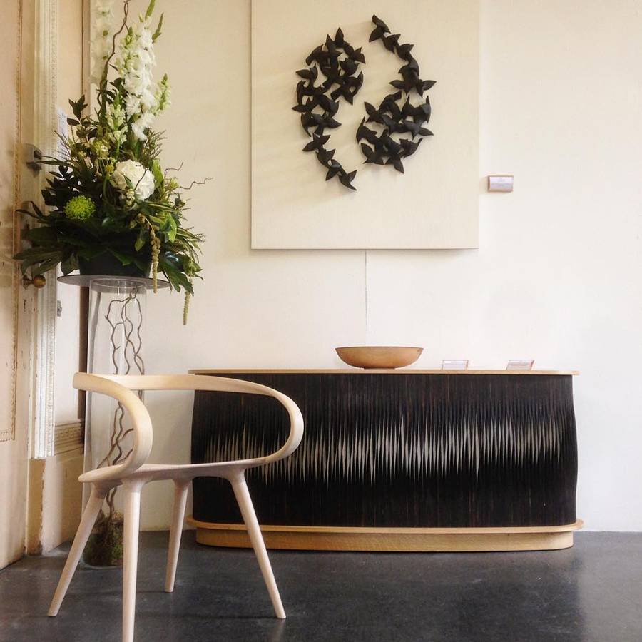 A curved fronted sideboard with a finely fluted scorched black front and a pale timber top and base, with a pale timber armchair in front, a pale wall panel of dark sculptural forms above and a tall floral arrangement beside it.