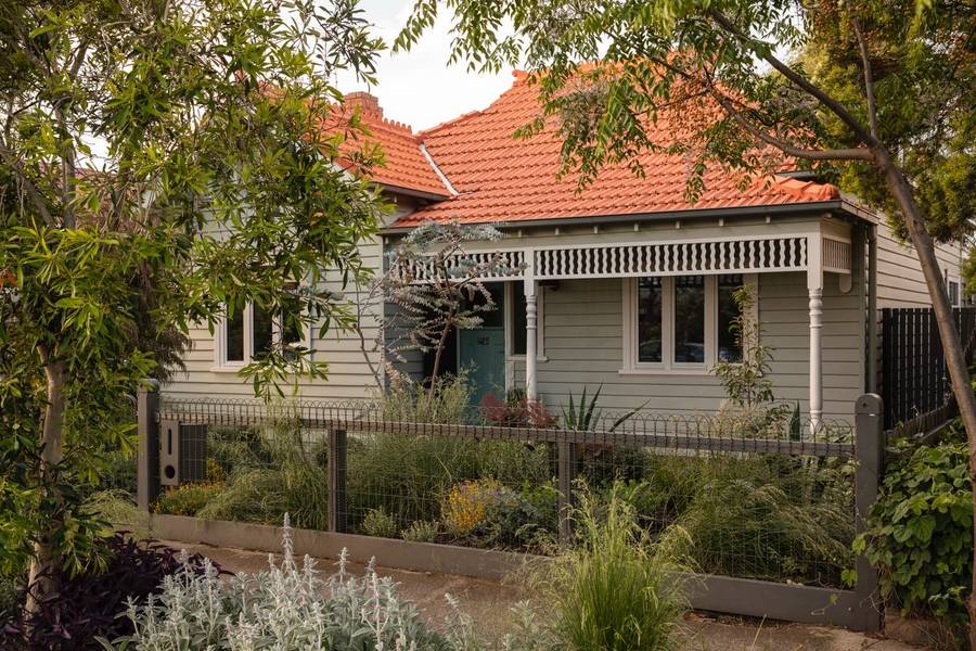 A weatherboard cottage with a terracotta tiled roof and a lace verandah, behind a densely planted front garden of grasses and silver foliage and a woven wire fence.