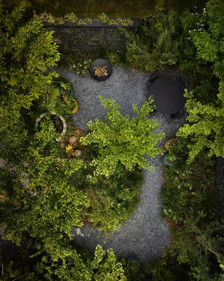 An overhead view of a small enclosed garden with a clearing of slate chippings and clay tile paving, a round black table, a fire bowl, ginkgo and pine planting and glazed pots.