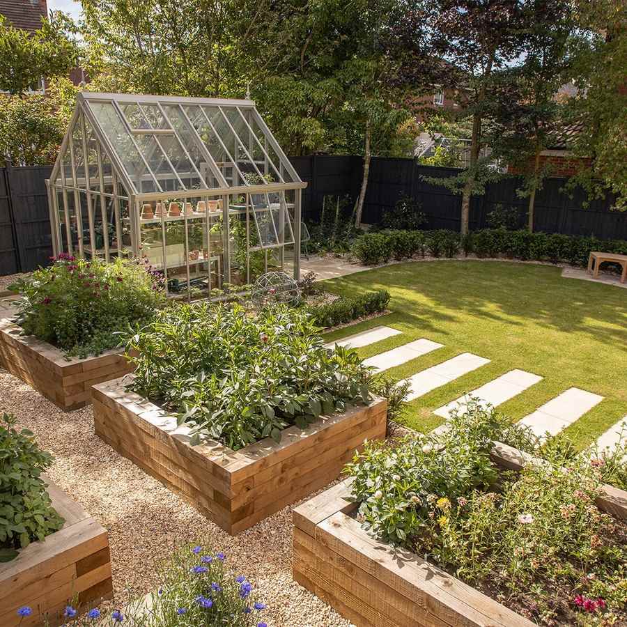 A back garden with a pale framed glasshouse, timber raised beds of vegetables and flowers, gravel paths, a lawn with stone slab steppers and a dark timber fence.
