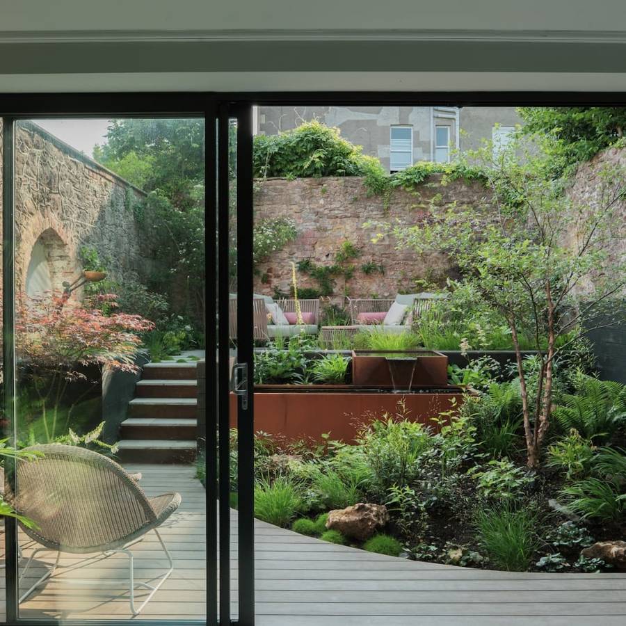 A small walled garden seen through open sliding glass doors, with a timber deck, a weathering steel raised bed with a water spout, a stone wall, an acer, ferns and seating on an upper terrace.