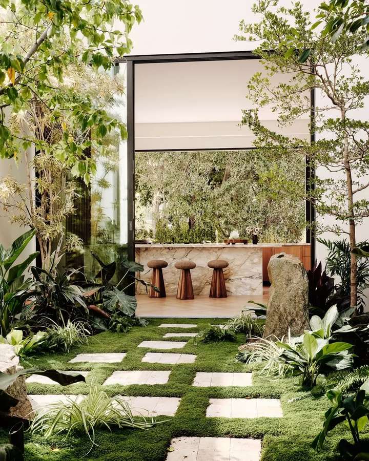 A courtyard of pale stone pavers set in green groundcover with boulders, ferns and broad leaved planting, looking through a full height opening to a kitchen with a marble island and timber stools.