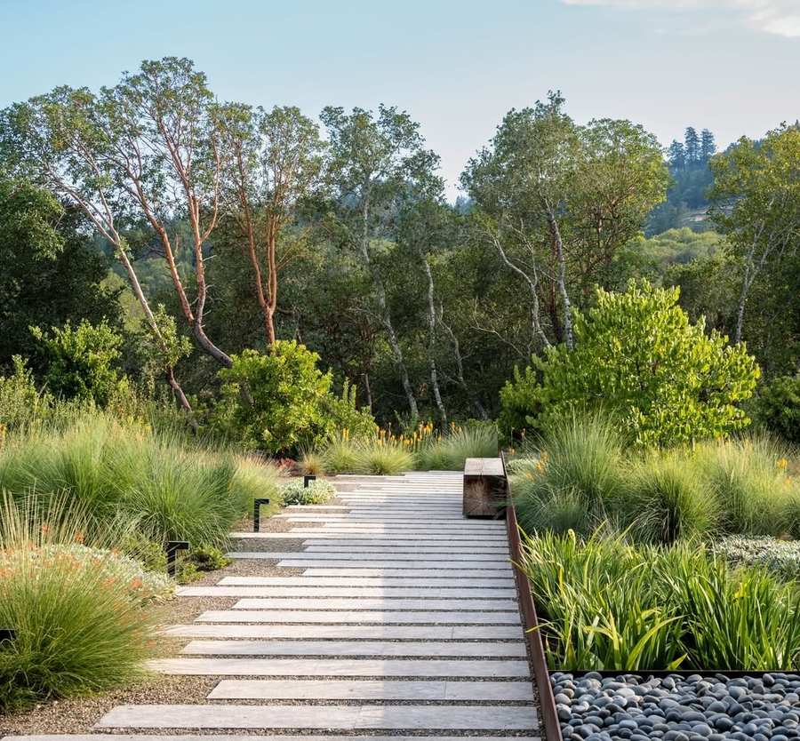 A broad path of long pale stone slabs set in gravel running between grasses and red hot pokers, with a timber bench beside a weathering steel edged bed of cobbles, and woodland and hills behind.