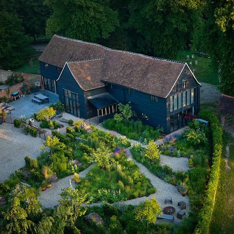 An overhead view of a black weatherboarded barn with a tiled roof, surrounded by a gravel garden of winding paths through beds of perennials, with a fire pit and a paved terrace.