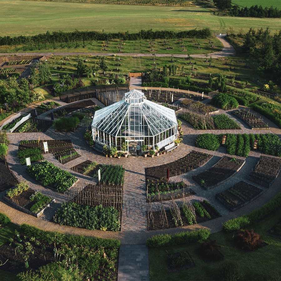 An overhead view of a circular kitchen garden with a white framed glasshouse at its centre, radiating raised beds of vegetables, gravel paths, and a young orchard and open fields beyond.
