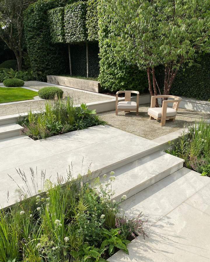 A garden of pale stone paving and steps with a gravel seating area, two pale timber armchairs with cream cushions under a multi stemmed tree, beds of grasses and white and pink flowering perennials, a clipped hedge and a lawn beyond.